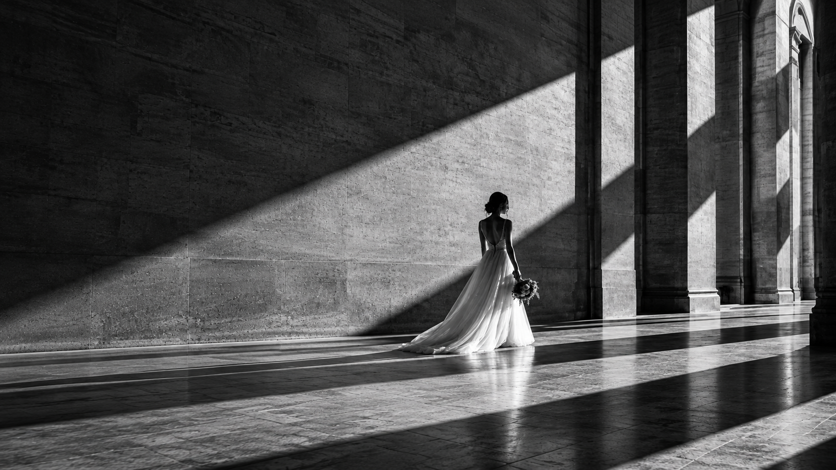 Sposa in abito bianco in una grande architettura di pietra, attraversata da fasci di luce e ombre geometriche in bianco e nero